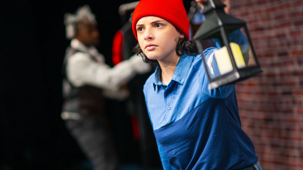 A young actor in a red hat and blue shirt holds up a lantern on a dark stage.