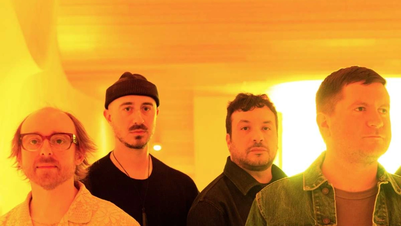 Four men in a band pose together in warm orange light.