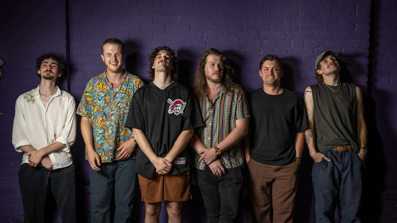 Six band members stand against a purple wall