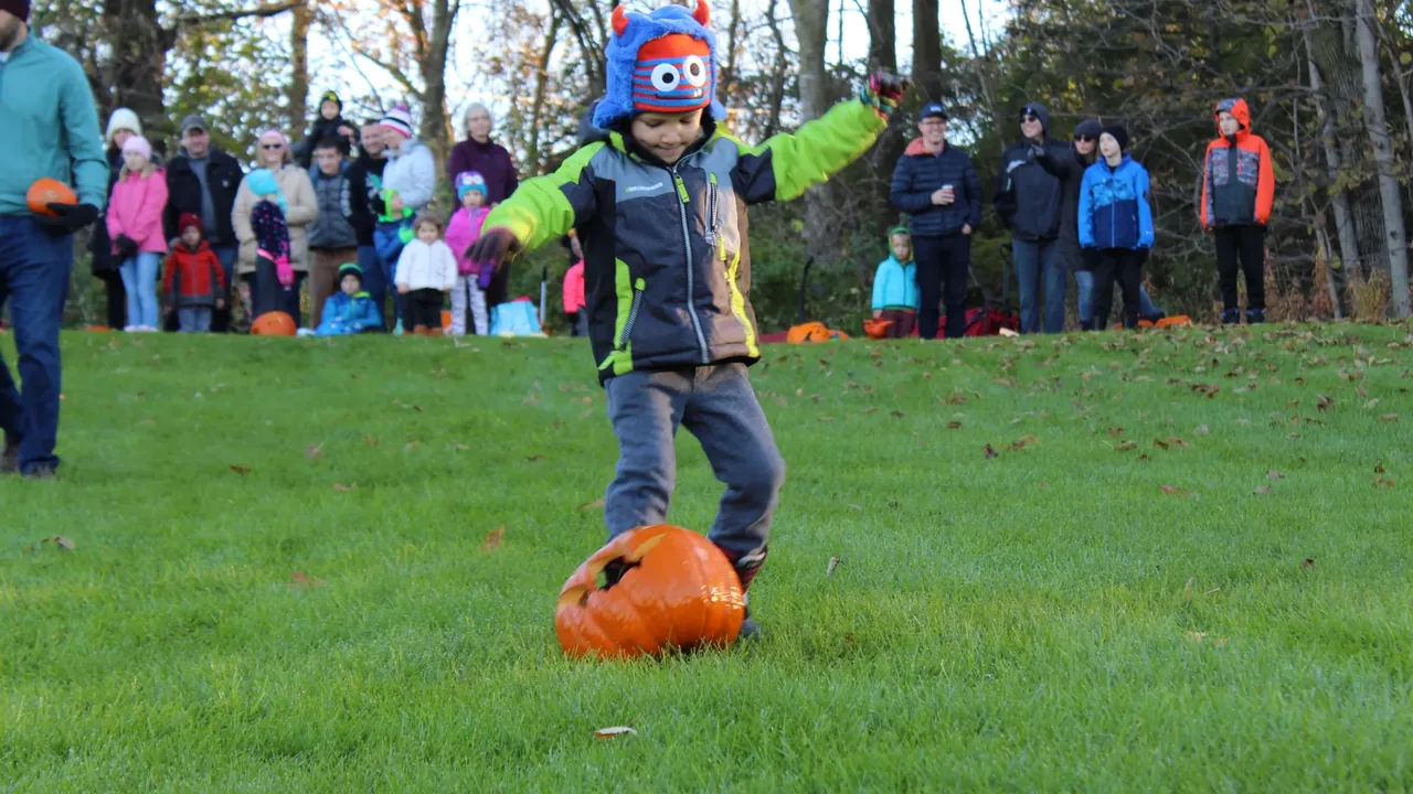A child in a costume kicks a pumpkin across a lawn as a crowd watches