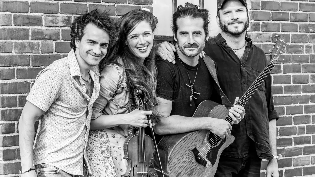 Four musicians pose against a brick wall in black and white