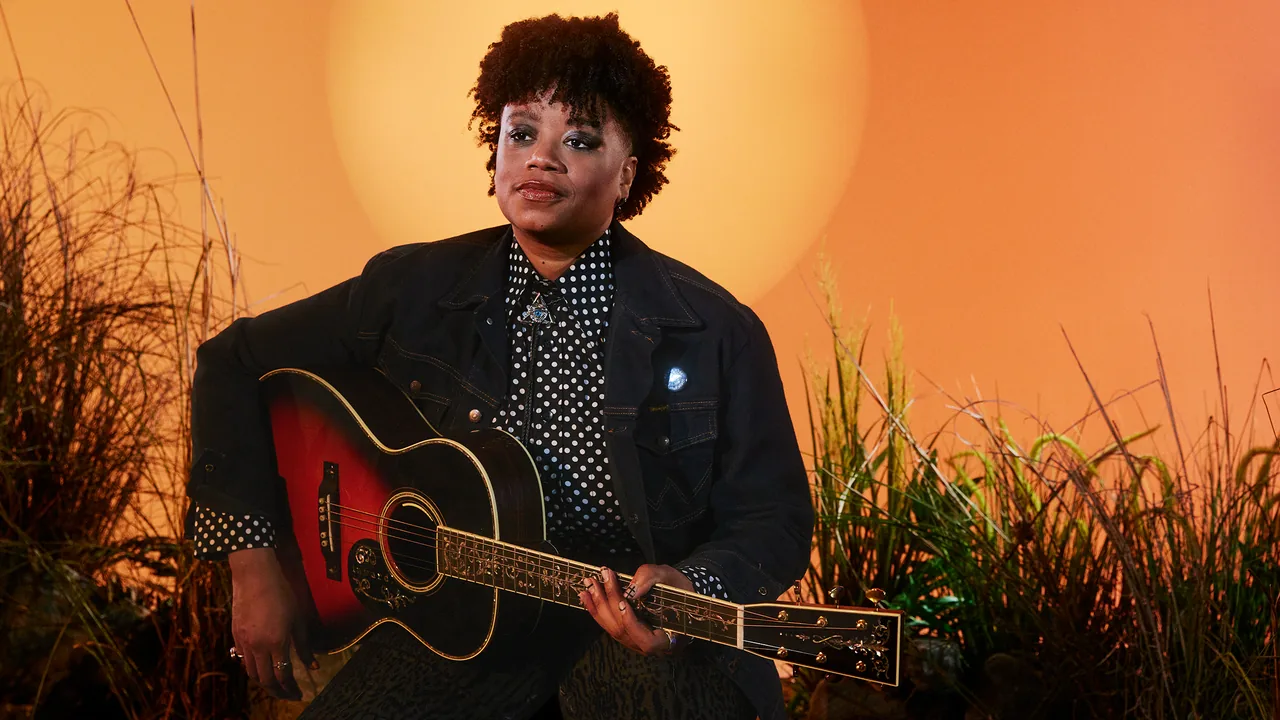 A woman with a guitar sits among tall grasses in warm light
