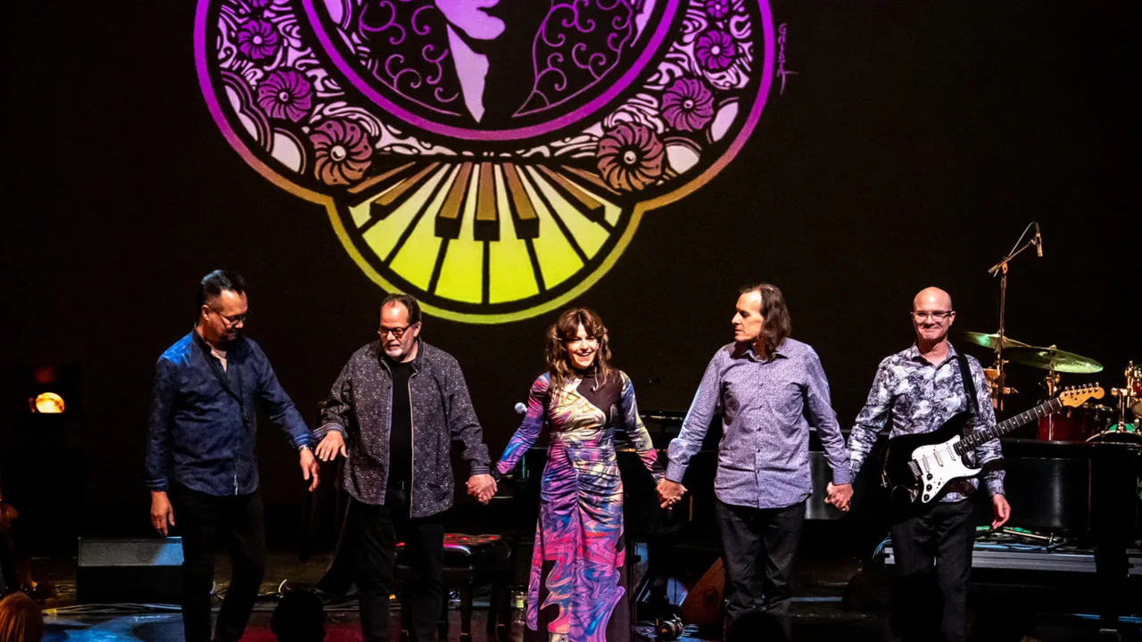 A five-piece band holds hands and bows on a colorfully lit stage