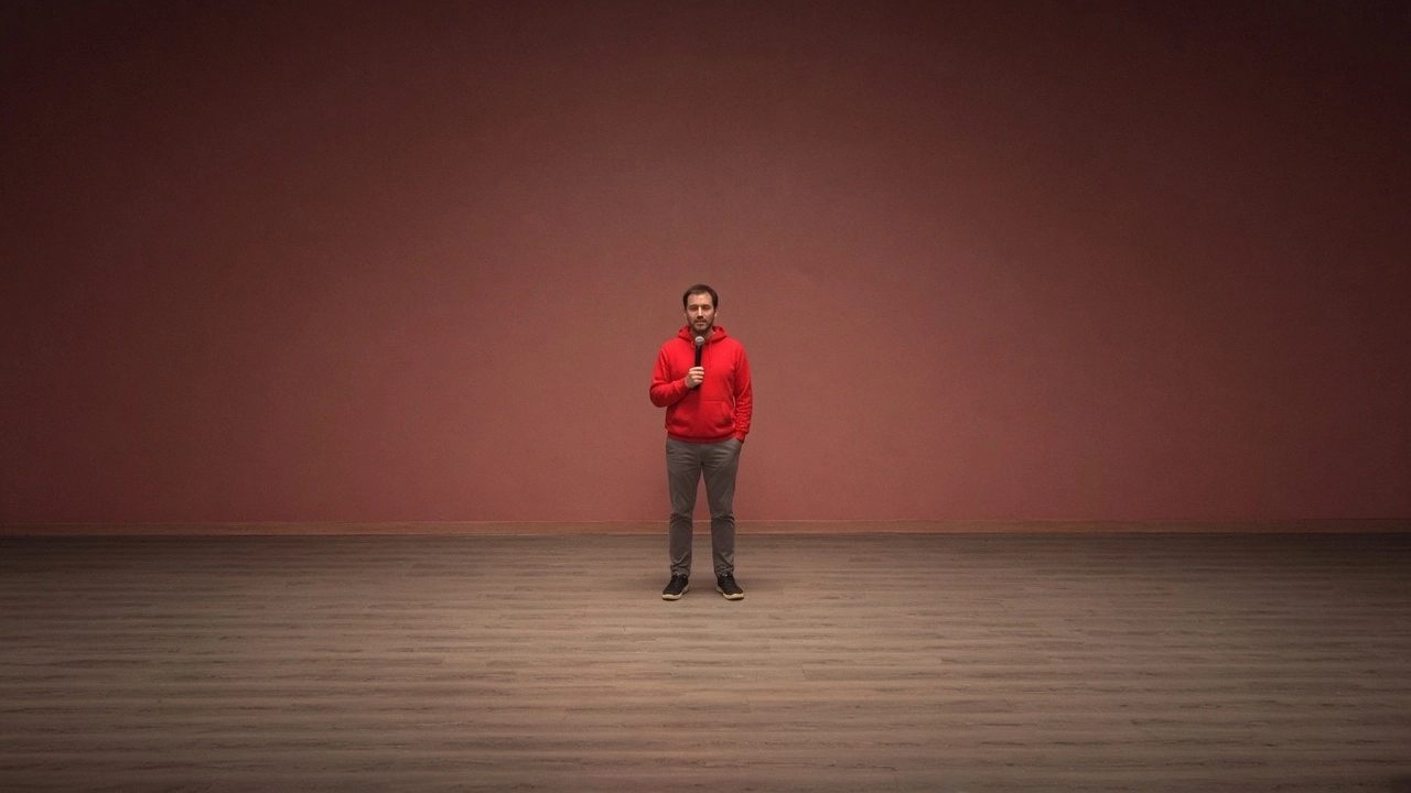 A comedian in a red jacket holds a microphone on an empty stage in front of a dark red wall.