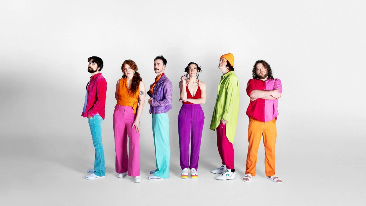 Six band members in brightly colored outfits pose against a white wall
