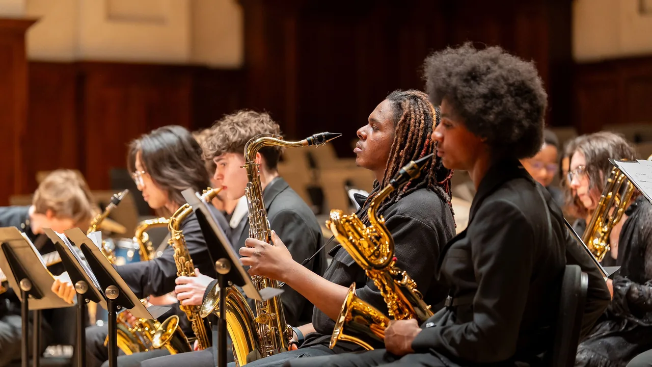 Young saxophone players in black perform in a jazz band on a wood-paneled stage.