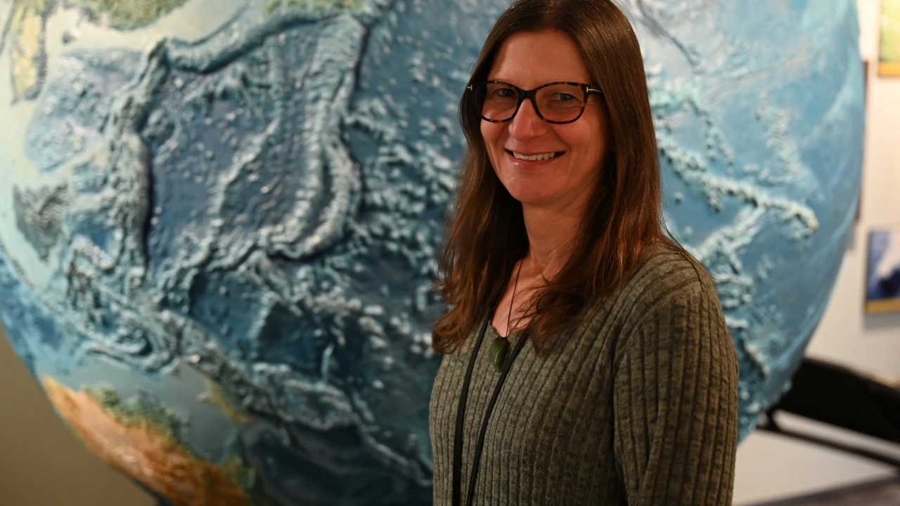 A smiling woman in glasses stands before a large globe