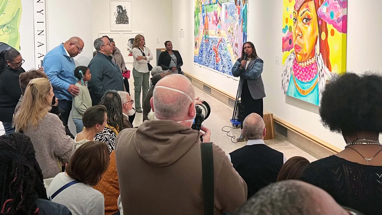 A crowd listens to a speaker in an art gallery with colorful paintings