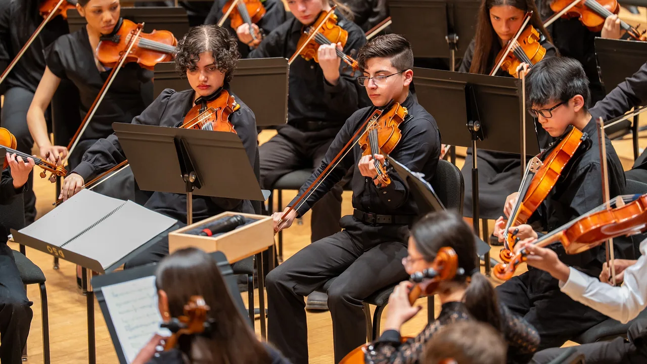 Young violinists in black play from music stands in a bright rehearsal hall.