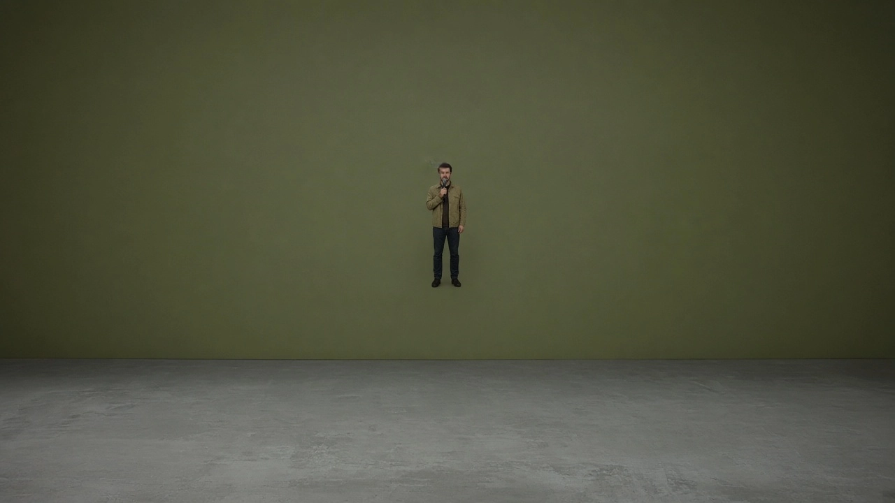 A comedian holds a microphone alone on a wide stage in front of an olive-green wall.