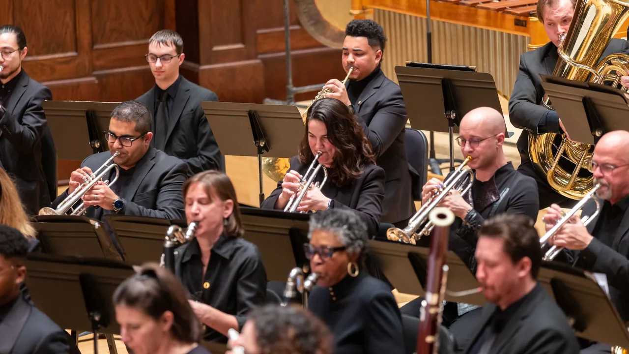 Brass and woodwind players in black concert dress perform seated in rows on stage.