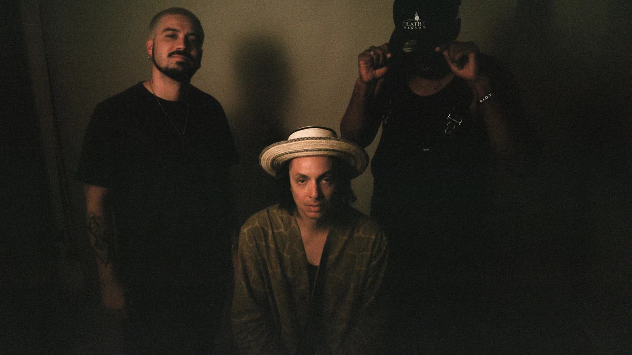 Three performers pose in a dim room, one in a straw hat