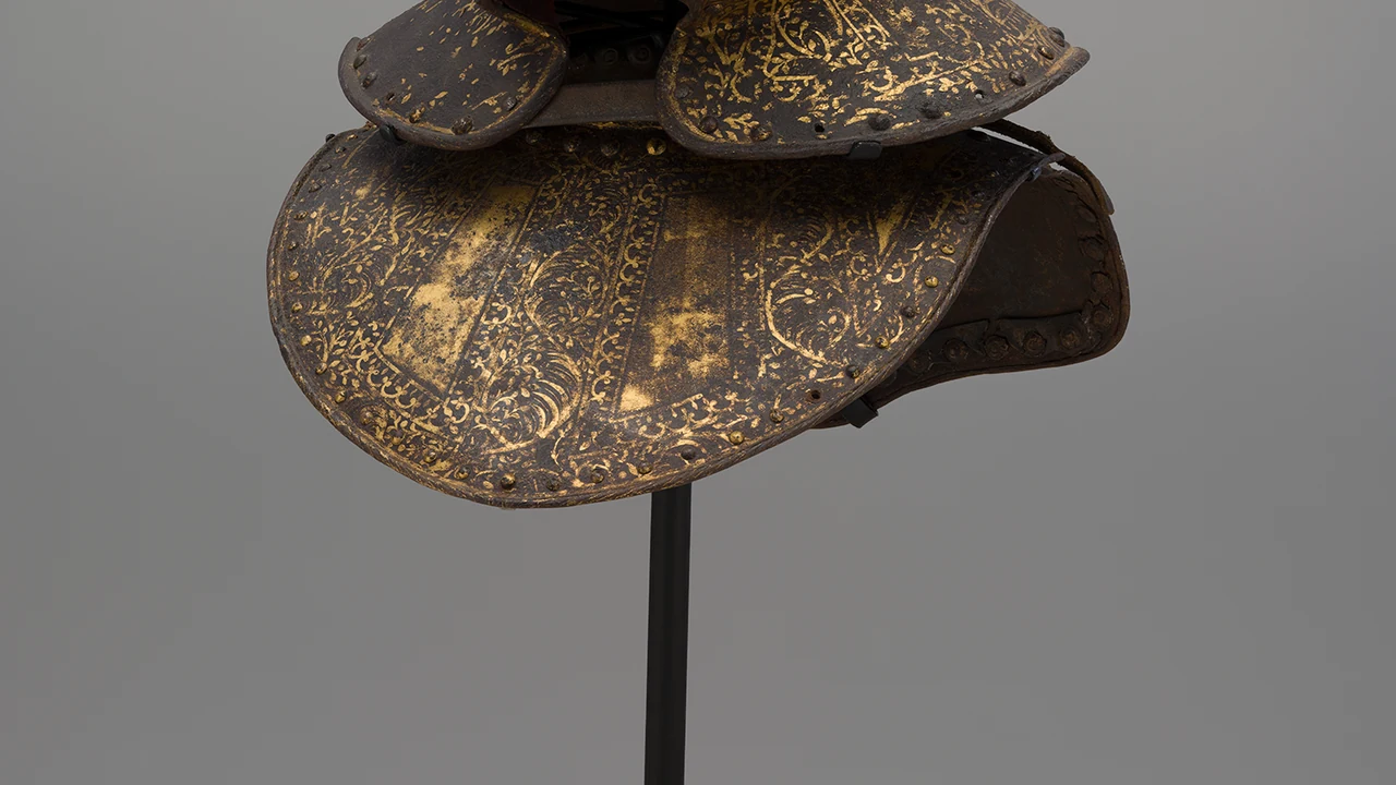 Ornate etched steel armor pauldron with gold scrollwork, displayed on a stand against a gray backdrop.