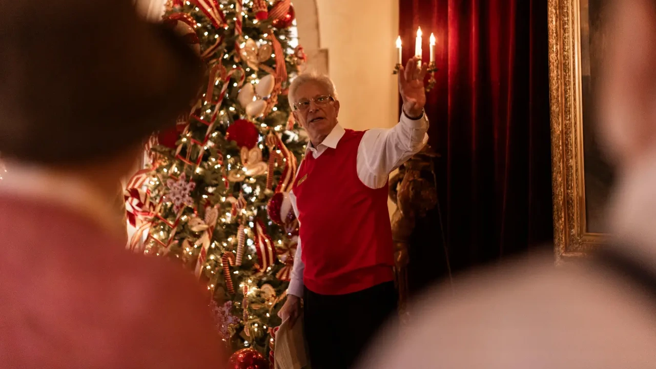A host in a red sweater raises a candelabra beside a decorated Christmas tree