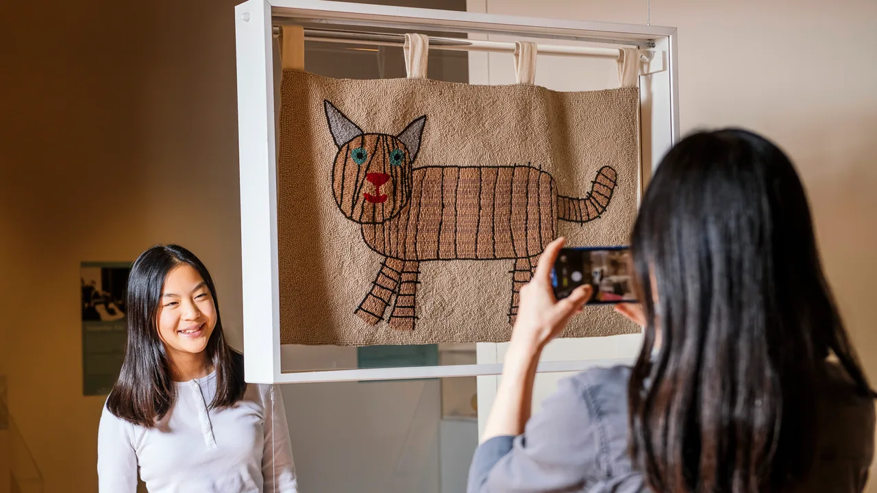 A young woman photographs a framed textile artwork in a gallery