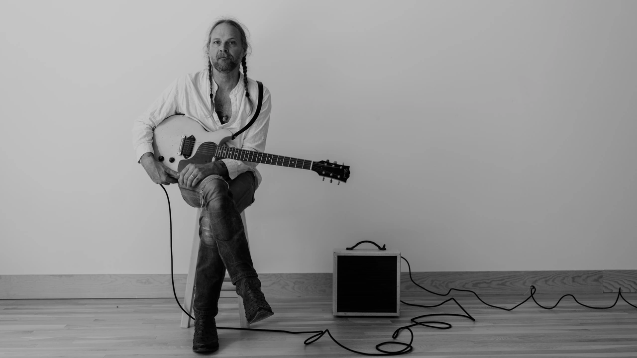 A long-haired guitarist sits on a stool beside an amp