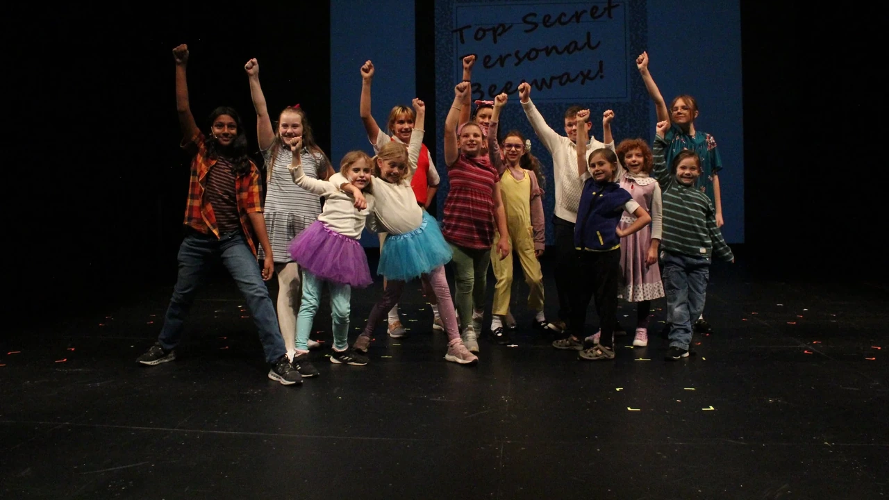 A young cast cheers with raised arms on a dark stage