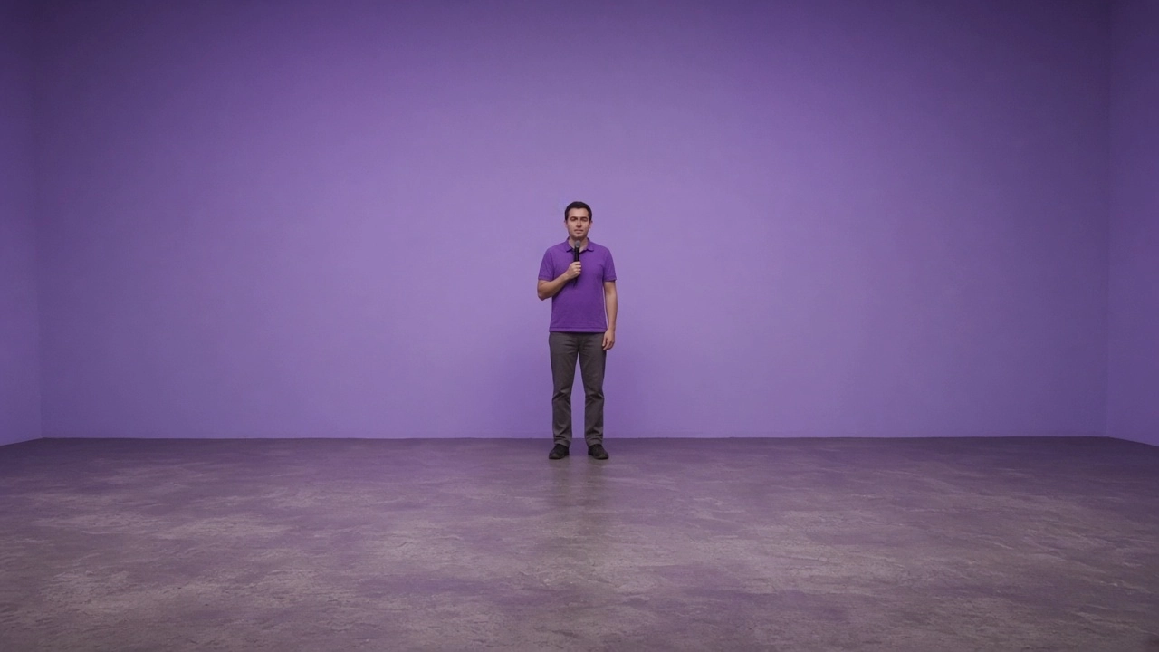 A comedian in a purple shirt holds a microphone on an empty stage in front of a lavender wall.