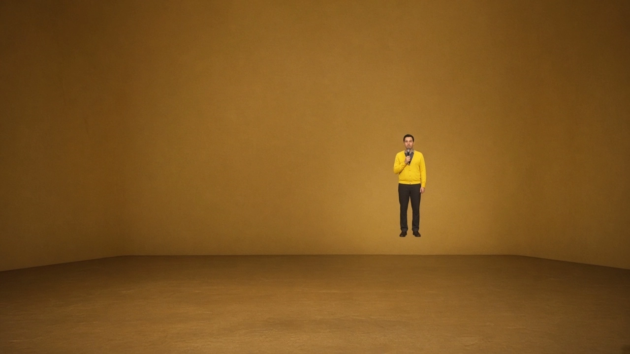 A comedian in a yellow shirt holds a microphone on an empty stage in front of a golden wall.