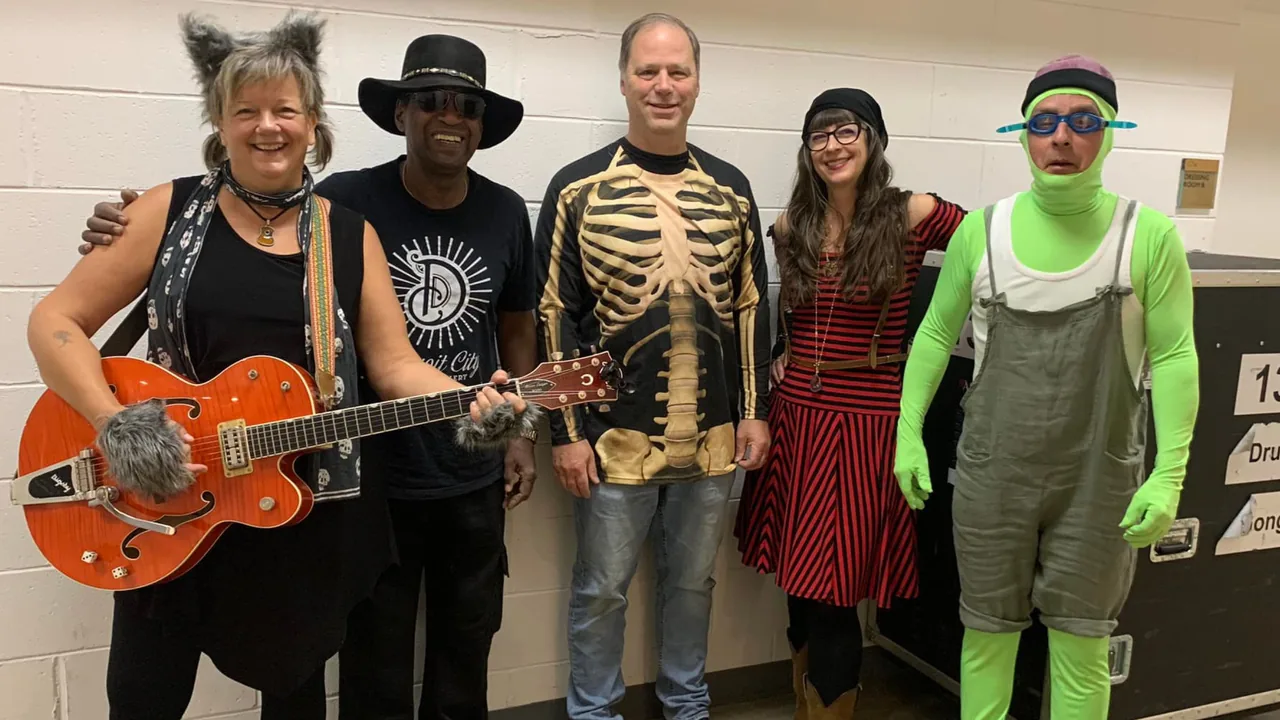 A costumed band poses with a guitarist in a skeleton shirt and a person in green