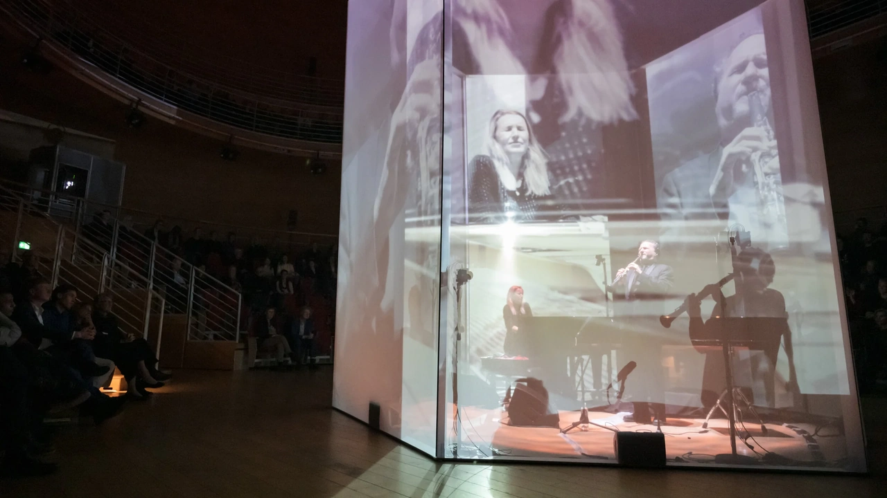 A performer at a piano on a stage under a large projection screen