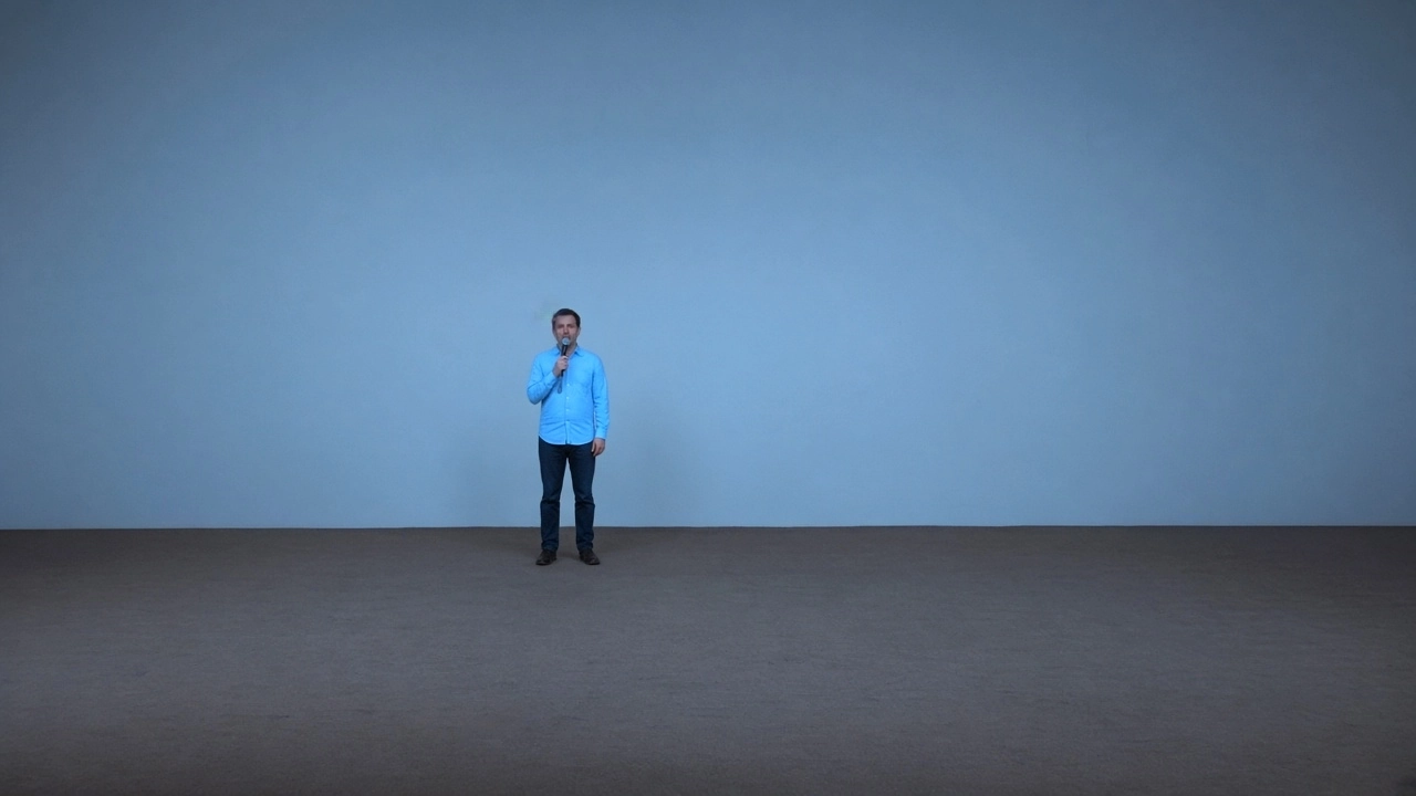 A comedian in a blue shirt holds a microphone on an empty stage in front of a pale blue wall.