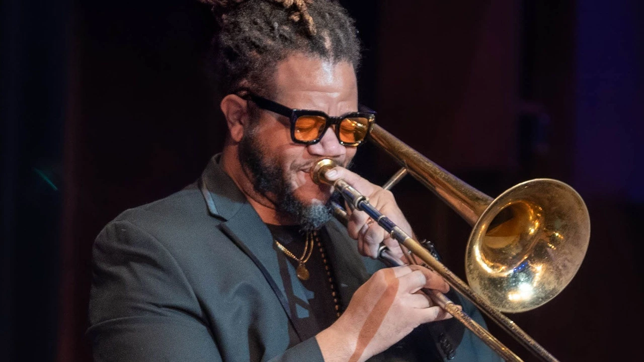 A trombonist in tinted glasses and a dark suit plays under stage lights.