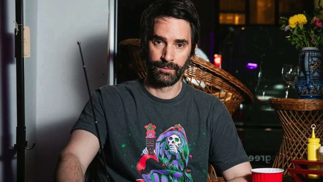 A bearded man in a graphic T-shirt sits in a cozy room