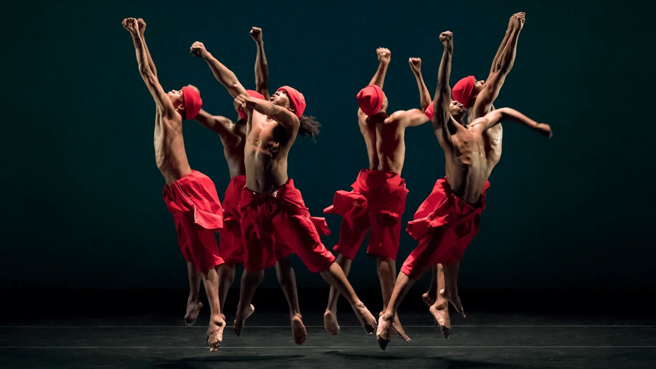 Dancers in red leap together across a dark stage