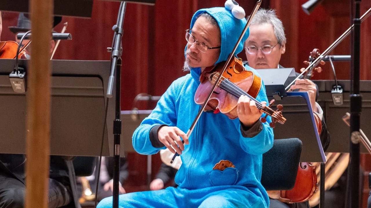 Orchestra violinist in a blue animal-costume onesie plays beside a fellow string player.