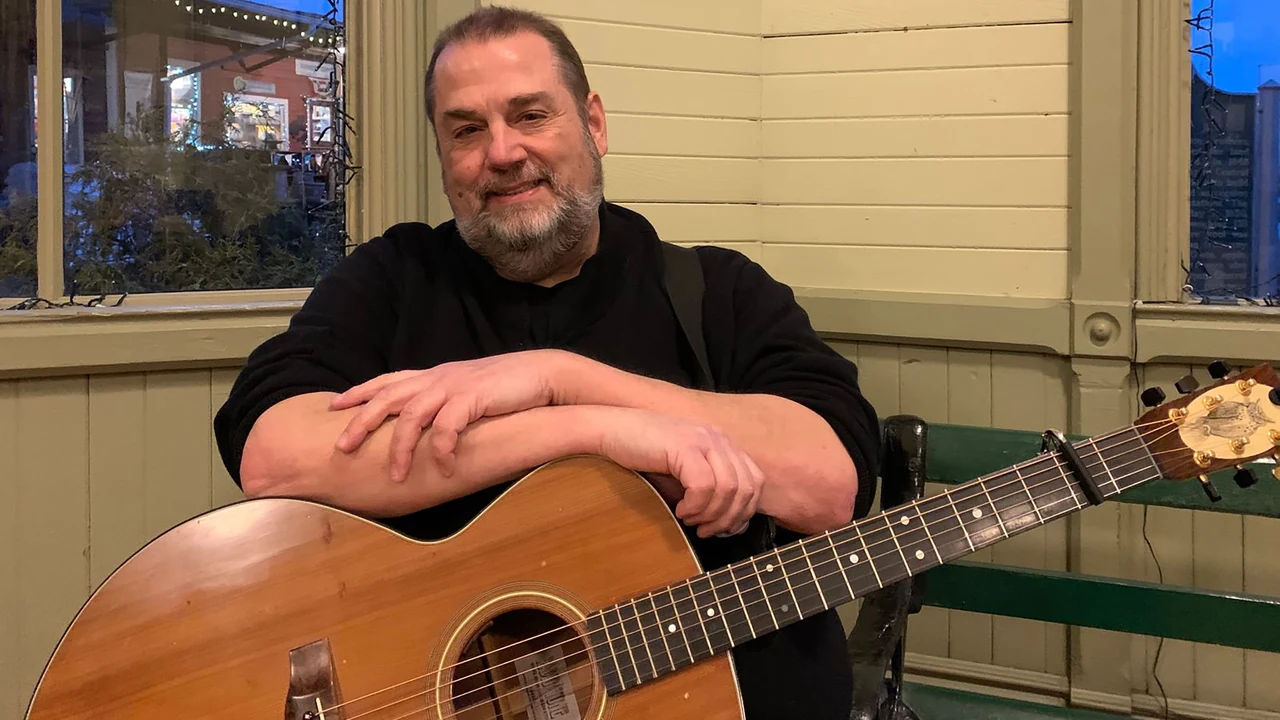A smiling guitarist sits with an acoustic guitar on a porch
