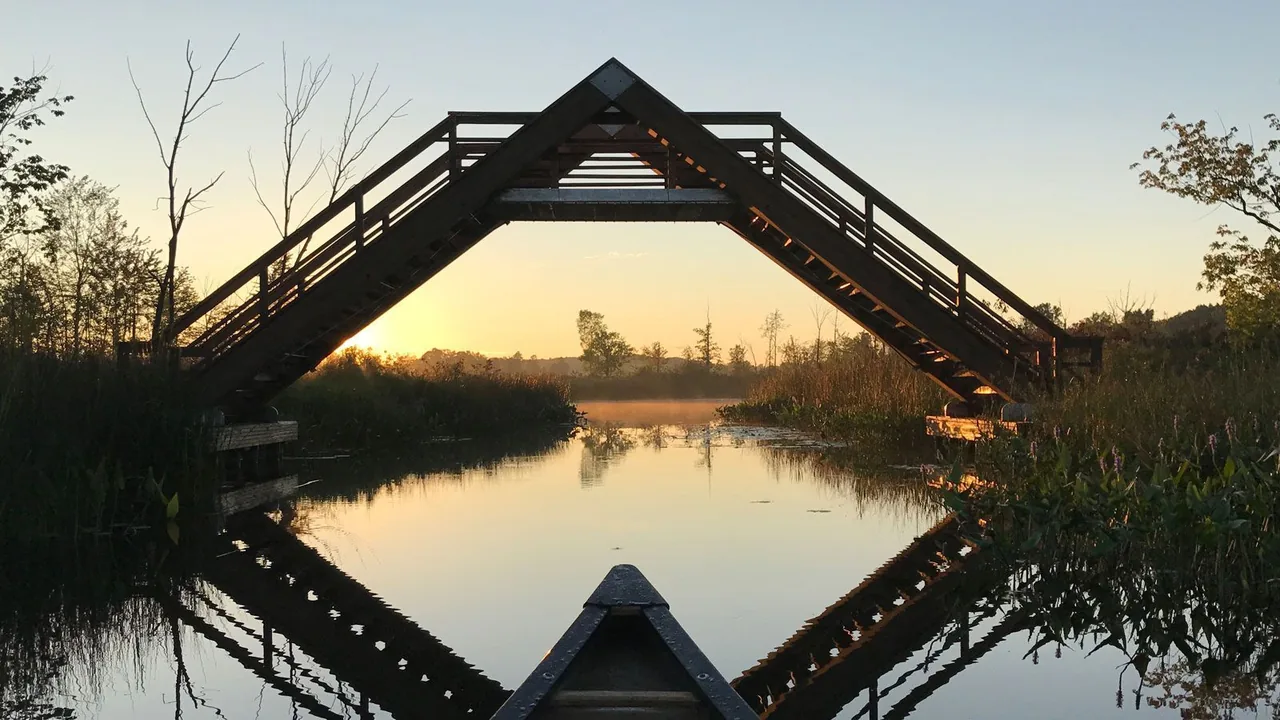A wooden footbridge arches over a still pond at sunset