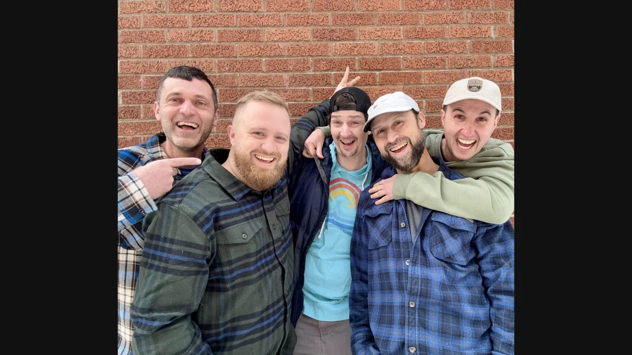 Five smiling men in flannel and caps pose by a brick wall