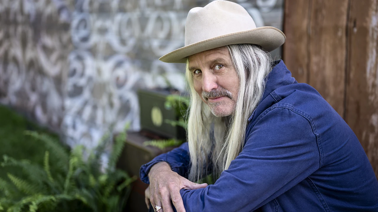 A long-haired man in a cowboy hat sits outdoors
