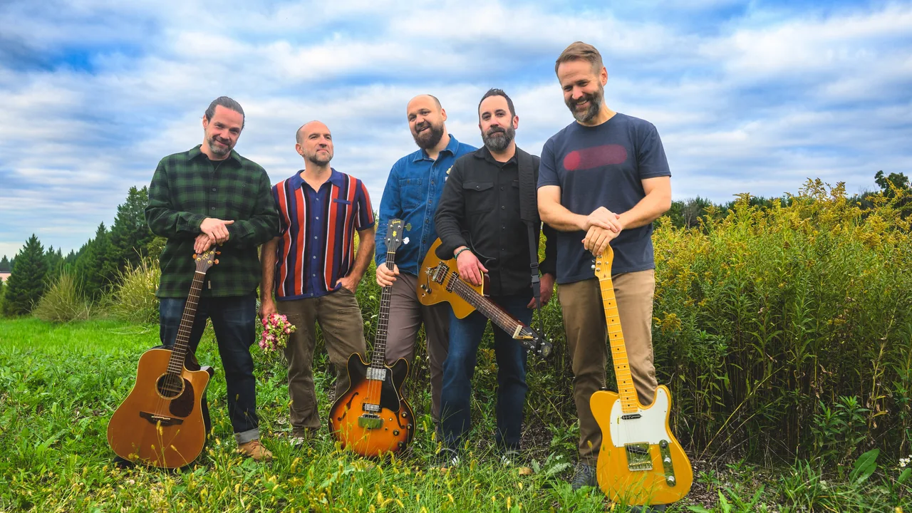 A five-piece band with guitars stands in a meadow
