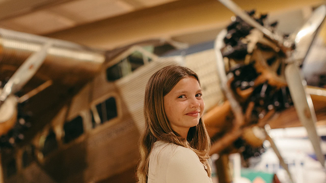 A girl smiles beside a vintage airplane in a museum hangar