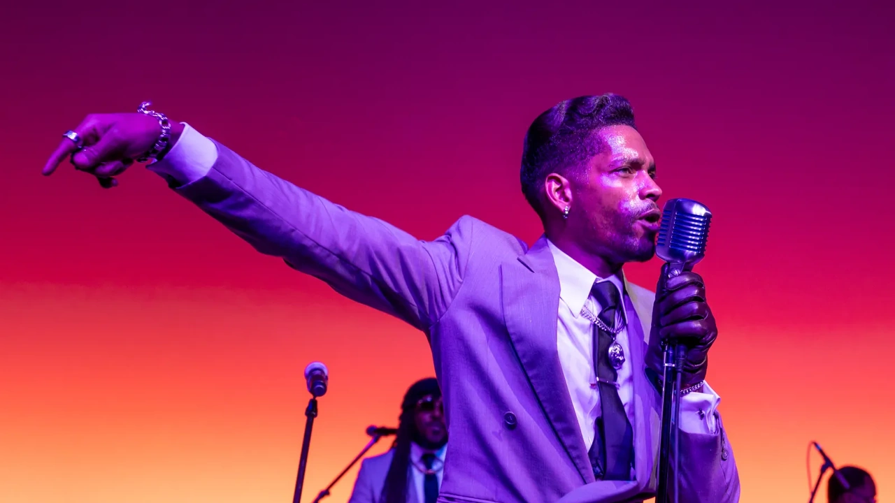 A singer in a purple suit points toward the crowd from a stage