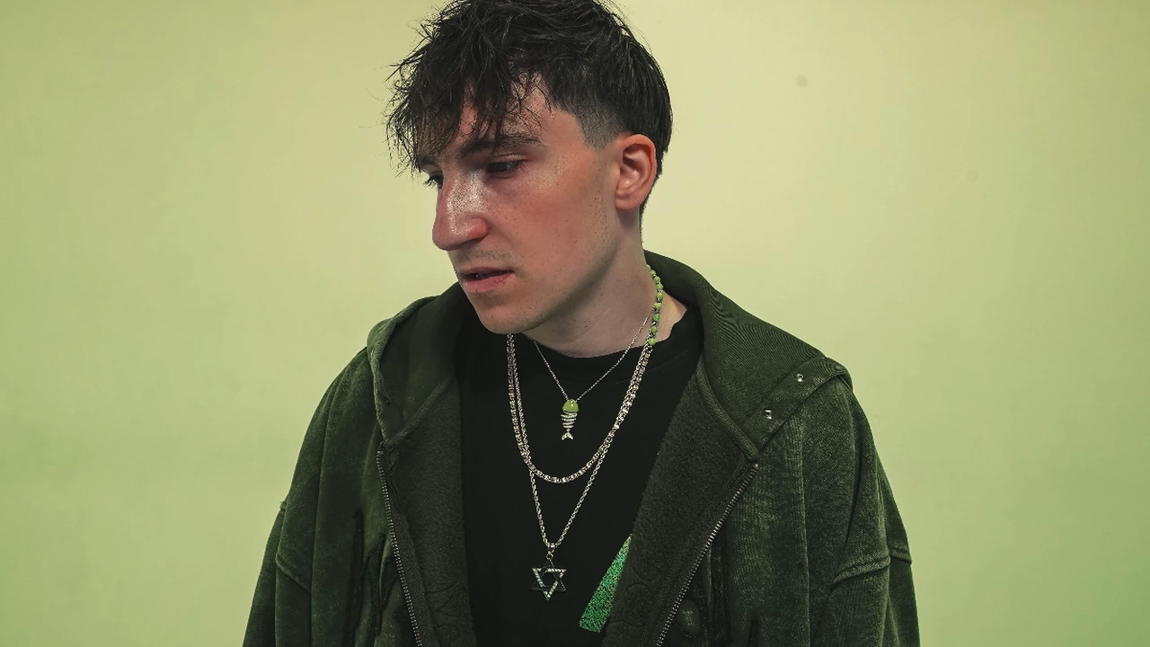 A young musician in a dark jacket and chains against a green wall
