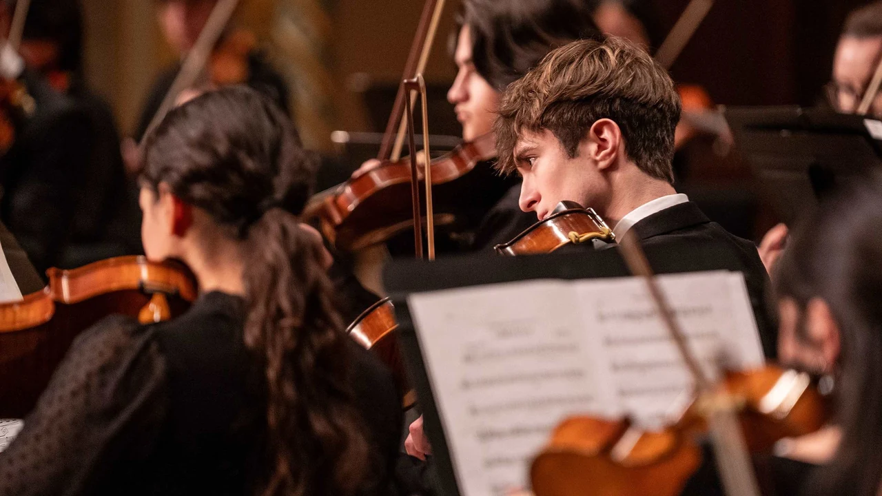 Young string players in black perform on stage with music on their stands.